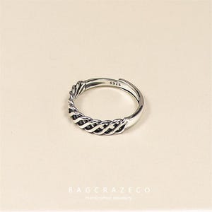 May include: A silver ring with a woven design. The ring is adjustable and stamped with "S925". This piece of jewellery is likely made of sterling silver. The ring is displayed against a neutral background.