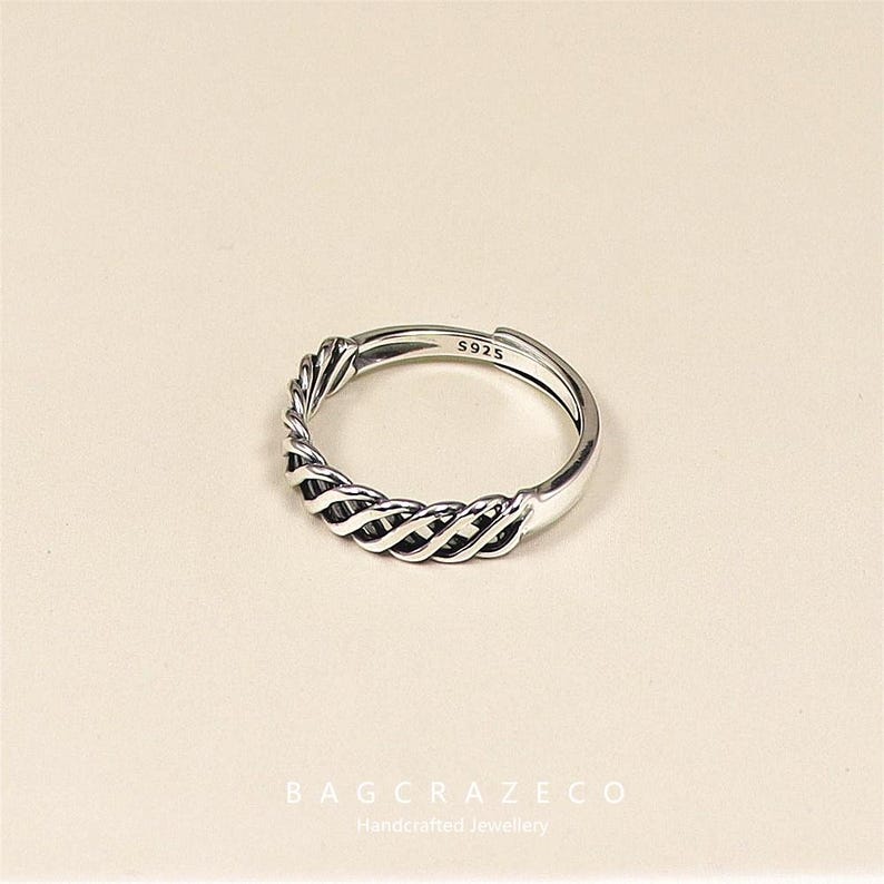 May include: A silver ring with a woven design. The ring is adjustable and stamped with "S925". This piece of jewellery is likely made of sterling silver. The ring is displayed against a neutral background.