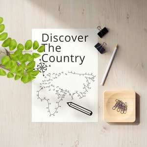 May include: A white sheet of paper with the text "Discover The Country" and a dot-to-dot map of a continent. A white pencil, two black binder clips, and a small wooden tray with paper clips are on a light wood surface. A green plant is in a white vase.
