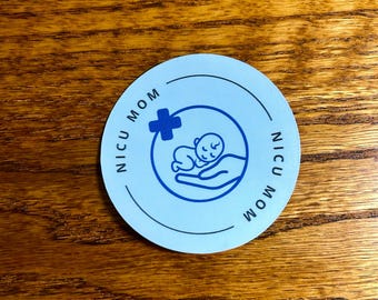 May include: A round, light blue sticker with the words "NICU MOM" in a circular pattern. The center features a blue cross and a baby cradled in hands. The sticker is on a wooden surface.