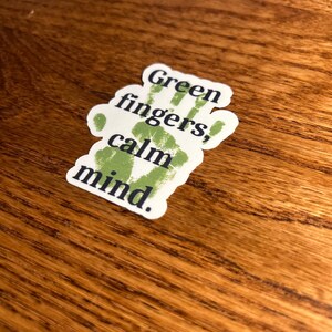 May include: A white sticker with the text "Green fingers, calm mind." The text is in a dark green color, with a handprint design. The sticker is on a wooden surface.