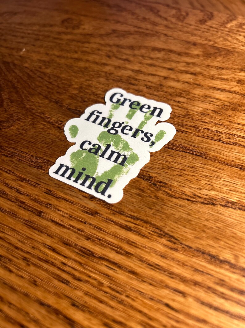May include: A white sticker with the text "Green fingers, calm mind." The text is in a dark green color, with a handprint design. The sticker is on a wooden surface.