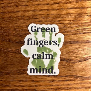 May include: A white sticker with the text "Green fingers, calm mind." The design includes a green handprint. The sticker is on a wooden surface.