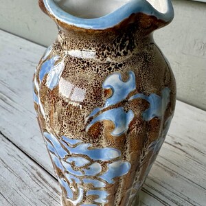 May include: A ceramic vase with a unique design. The vase features a brown and tan speckled background with light blue leaf-like patterns. The rim is a light blue color, and the interior is white.