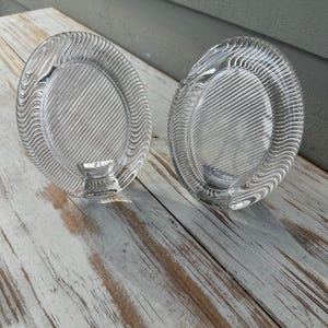 May include: Two clear glass candle holders with an oval shape. Each holder features a textured surface with a pattern of diagonal lines and wavy ridges along the outer rim, surrounding a smooth central area with a small circular base for a candle.