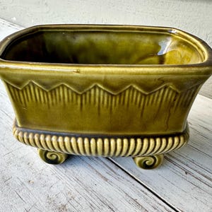 May include: A rectangular, olive-green ceramic planter with a decorative zigzag pattern and vertical lines. The planter has a curved top and rests on four scrolled feet. The planter is designed for indoor or outdoor use.