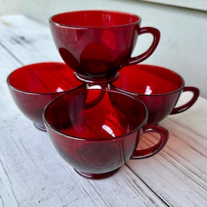 May include: Four ruby red glass teacups with handles are stacked on a white wooden surface. The cups have a rounded shape and a smooth, glossy finish. The cups are arranged in a pyramid shape.