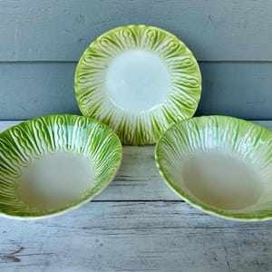 May include: Three ceramic serving dishes with a lettuce leaf design. The set includes two oval bowls and one round plate. The rims are a bright green, and the interiors are a creamy white. The dishes are arranged on a light-colored wooden surface.
