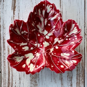 May include: A leaf-shaped ceramic bowl featuring a glossy finish with a deep red base color and irregular cream-colored splotches. The surface displays embossed veins radiating from the center, mimicking the natural structure of a plant leaf.