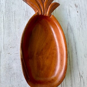 May include: A wooden pineapple-shaped serving dish. The dish is carved from a light brown wood and features a detailed pineapple top. The oval-shaped bowl is smooth and has a glossy finish. The dish is designed for serving snacks or small items.