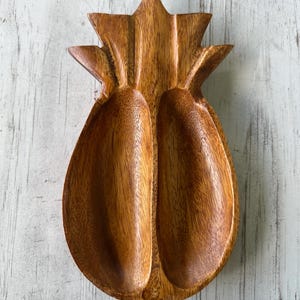 May include: A wooden pineapple-shaped serving dish with two compartments. The dish is made of a light brown wood and has a natural grain pattern. The top of the dish is shaped like a pineapple crown.