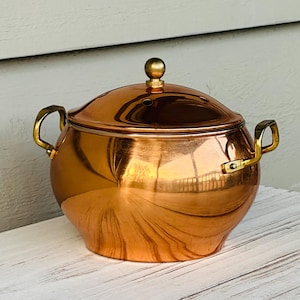 May include: A copper pot with a lid and brass handles. The pot has a rounded body and a shiny, reflective surface. The lid has a small brass knob and several small holes. The handles are curved and attached to the sides of the pot.