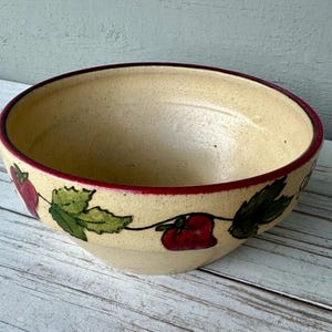 May include: A beige ceramic bowl with a red rim and a decorative pattern of red strawberries and green leaves. The bowl has a wide, shallow shape and is set against a light-colored wooden surface.
