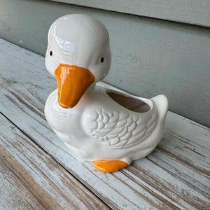 May include: A white ceramic duck planter with a glossy finish, featuring a bright orange beak and orange feet. The duck has molded feather details on its wings and a hollow opening on its back for holding small items or plants.