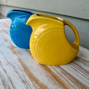 May include: Two ceramic pitchers with a glossy finish, one in a bright yellow and one in a vibrant blue. Each pitcher features a rounded body, a curved handle, and a wide pouring spout, with concentric ring detailing embossed on the side surfaces.