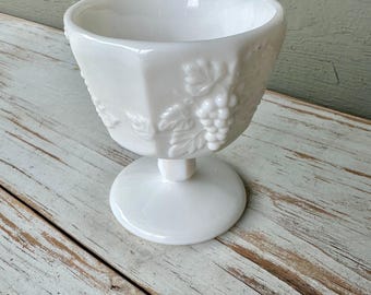 May include: White, vintage-style glass dessert dish with a pedestal base. The bowl has a hexagonal shape and is decorated with a grape and leaf motif. The dish is sitting on a distressed, white-painted wooden surface.