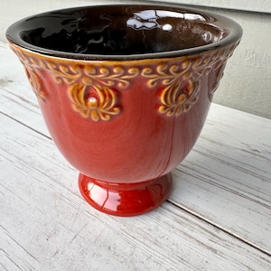 May include: A red ceramic planter with a decorative floral pattern and a dark brown interior. The planter has a pedestal base and a wide, slightly flared rim. The exterior has a crackled glaze finish.