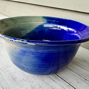 May include: A large, handmade ceramic bowl with a wide rim. The bowl features a gradient of colors, transitioning from a deep blue at the base to a lighter blue and green at the top. The bowl is sitting on a white wooden surface.