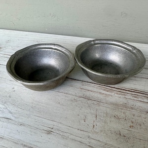 May include: Two antique pewter bowls with a scalloped rim. The bowls have a slightly textured surface and a dark, metallic gray color. They are placed on a weathered, light-colored wooden surface. The bowls are empty and appear to be vintage.