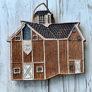 May include: A brown ceramic barn-shaped wall hanging with a dark blue roof and white accents. The barn features multiple windows and doors, and a small cupola. A leather cord is attached for hanging.