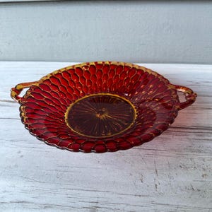 May include: A ruby red glass dish with a textured honeycomb pattern and two small handles. The center of the dish has a starburst design. The rim and handles have a gold trim. This decorative dish is likely vintage.