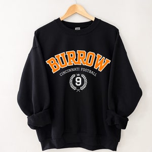 Może przedstawiać: Czarna bluza z pomarańczowym napisem "Burrow Cincinnati Football" z białym wieńcem i cyfrą 9 w środku.