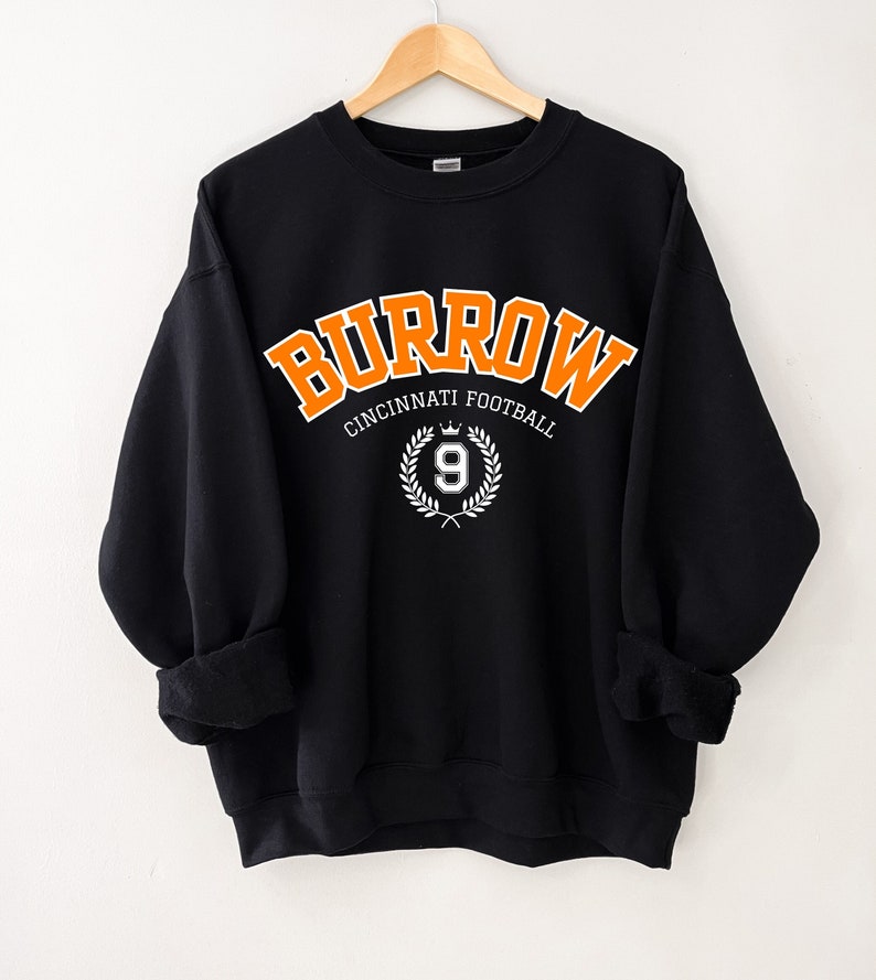 Może przedstawiać: Czarna bluza z pomarańczowym napisem "Burrow Cincinnati Football" z białym wieńcem i cyfrą 9 w środku.