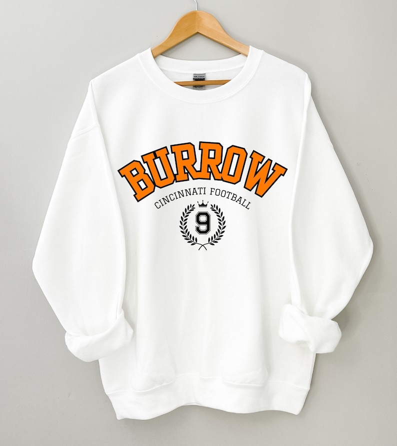 Może przedstawiać: Biała bluza z pomarańczowym napisem "Burrow Cincinnati Football" z czarno-białym wieńcem i cyfrą 9 w środku.