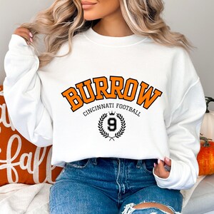 Może przedstawiać: Biała bluza z pomarańczowym napisem "Burrow Cincinnati Football" z czarną koroną i wieńcem laurowym z numerem 9 w środku.