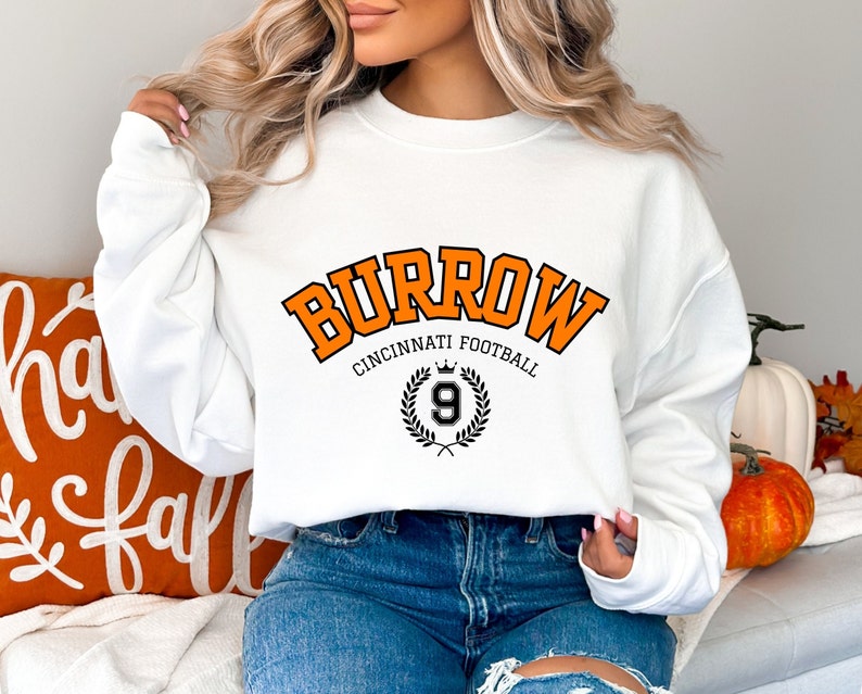 Może przedstawiać: Biała bluza z pomarańczowym napisem "Burrow Cincinnati Football" z czarną koroną i wieńcem laurowym z numerem 9 w środku.