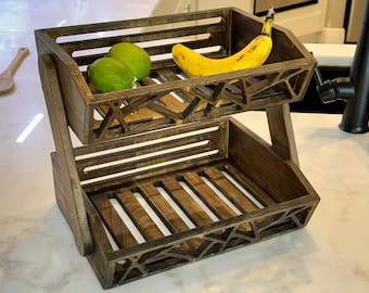 May include: A two-tiered wooden fruit basket with a dark brown finish. The top tier holds two green limes and a yellow banana. The basket has a geometric design on the sides and is suitable for kitchen or dining room use.