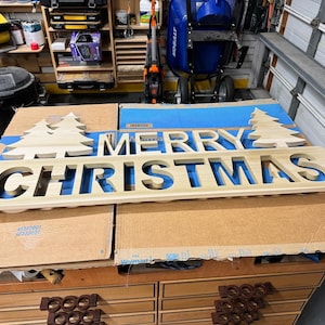 Może przedstawiać: Drewniany znak z wyciętymi słowami "Merry Christmas". Znak jest pomalowany na jasnobrązowy kolor i ma dwa choinki po obu stronach słów.