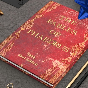 Könnte beinhalten: Ein rotes Buch mit dem Titel "The Fables of Phaedrus" mit goldener Schrift und dekorativen Bordüren. Das Buch trägt die Aufschrift "1886 First Edition". Eine blaue Schleife, goldene und schwarze Bleistifte sowie goldene Büroklammern sind ebenfalls vorhanden.