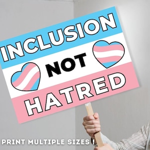 Puede incluir: Un cartel rosa, azul y blanco con el texto "Inclusion Not Hatred" y dos corazones de orgullo transgénero. El cartel está sostenido por la mano de una persona.