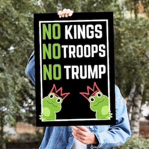 May include: Black sign with white border, held in front of a person. The sign reads "NO KINGS, NO TROOPS, NO TRUMP" in green and white text. Two cartoon frogs wearing crowns are at the bottom.