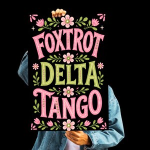 May include: A black sign with the words "Foxtrot Delta Tango" in pink and green, surrounded by floral designs. The sign is held up by a person wearing a denim jacket against a black background.