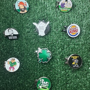 May include: Assortment of nine golf ball markers with various designs and sayings. The markers are round and feature a variety of colors, including green, red, blue, white, and black. The sayings on the markers include "That's what she said", "The price is wrong", "Play loose", "Bogey King", "I'd tap that", "This is how I roll", and more.