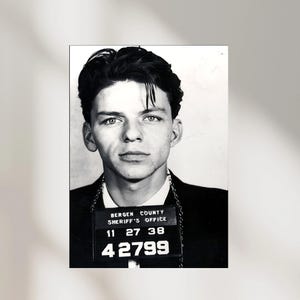 May include: Black and white mugshot of a man with dark hair and a serious expression. He is wearing a dark suit and a chain. A sign around his neck reads "Bergen County Sheriff's Office" with the numbers "11 27 38" and "42799".