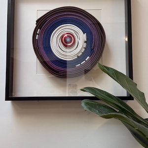 May include: Framed artwork featuring a circular design made of layered martial arts belts in shades of brown, purple, blue, and white. A Captain America shield is centered. The artwork is displayed in a black frame.