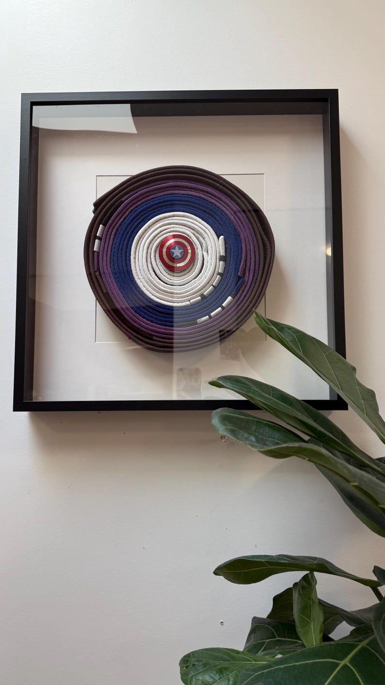 May include: Framed artwork featuring a circular design made of layered martial arts belts in shades of brown, purple, blue, and white. A Captain America shield is centered. The artwork is displayed in a black frame.