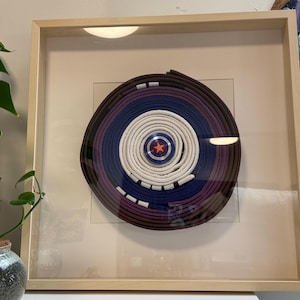 May include: Framed artwork featuring a circular design with concentric layers of dark brown, purple, blue, and white. A red star is at the center. The artwork is displayed in a light wood frame.