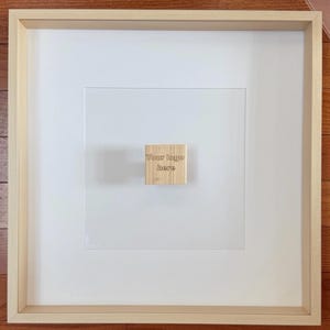 May include: A framed artwork with a light wood frame and a white mat. A small, square wooden block with the text "Your logo here" is affixed to the center of the artwork. The artwork is displayed on a light wood surface.