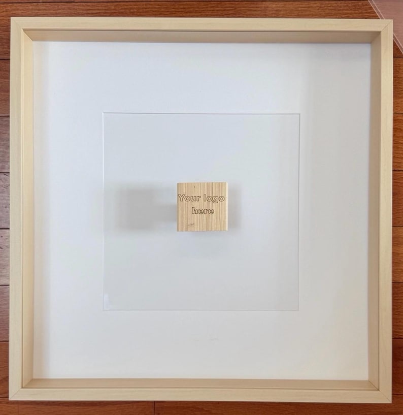 May include: A framed artwork with a light wood frame and a white mat. A small, square wooden block with the text "Your logo here" is affixed to the center of the artwork. The artwork is displayed on a light wood surface.