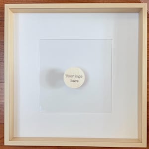 May include: A framed artwork with a light wood frame and a white mat. A small, round wooden piece with the text "Your logo here" is affixed to the center of the artwork. The artwork is displayed on a wooden surface.