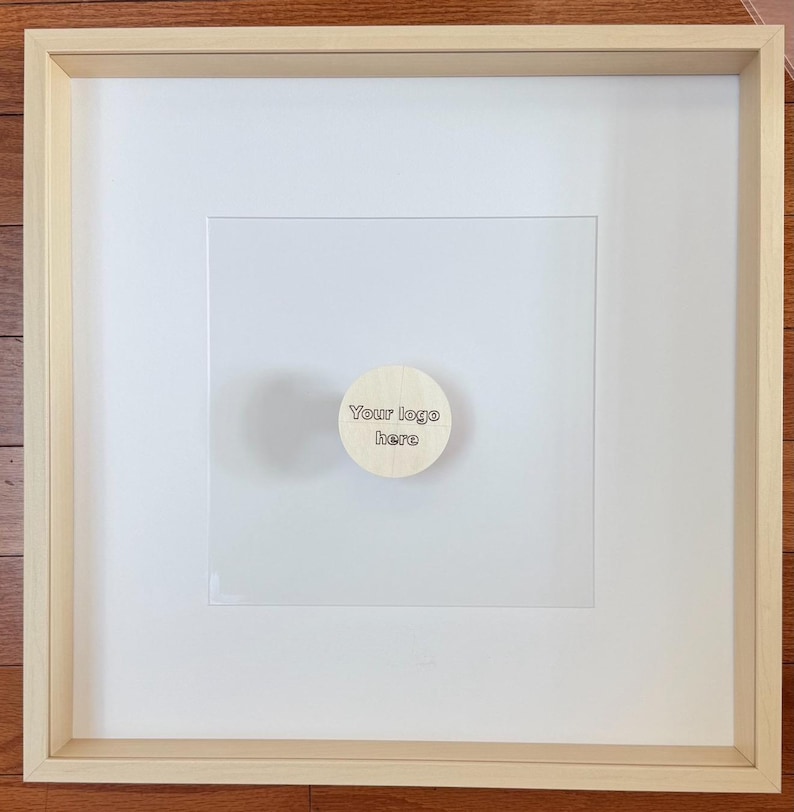May include: A framed artwork with a light wood frame and a white mat. A small, round wooden piece with the text "Your logo here" is affixed to the center of the artwork. The artwork is displayed on a wooden surface.