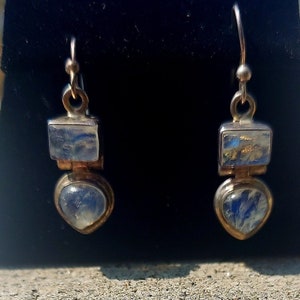 May include: A pair of silver-toned earrings with a rectangular top and a heart-shaped bottom. Each earring features a blue and white iridescent stone. The earrings are displayed against a black background.