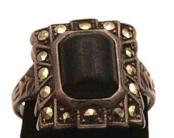 May include: A silver ring with a black stone set in a square frame surrounded by smaller stones. The ring is on a black velvet display stand.