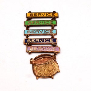 May include: A vintage service award pin with five rectangular bars in yellow, green, turquoise, navy, and lavender, each with the word "SERVICE" in gold lettering. A gold-toned cauldron-shaped emblem hangs below.