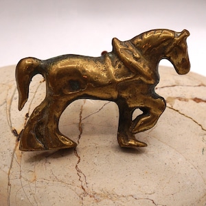 May include: A small, gold-colored metal horse figurine with a saddle. The horse is standing on a light brown marble surface.