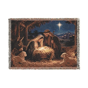 May include: A rectangular woven throw blanket featuring a detailed nativity scene. The tapestry depicts a central manger with figures, a donkey, and two lambs in a stable setting under a bright star, finished with a decorative fringed edge.